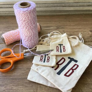 K&ouml;nnte beinhalten: Vier kleine wei&szlig;e Stoffbeutel mit Kordelzug. Die Beutel sind &uuml;bereinander gestapelt. Der gr&ouml;&szlig;te Beutel tr&auml;gt die Buchstaben "RTB" in Rot mit einem schwarzen Hammer-Motiv. Die kleineren Beutel haben das gleiche Design, sind aber kleiner. Eine orangefarbene Schere und zwei Garnrollen befinden sich im Hintergrund.