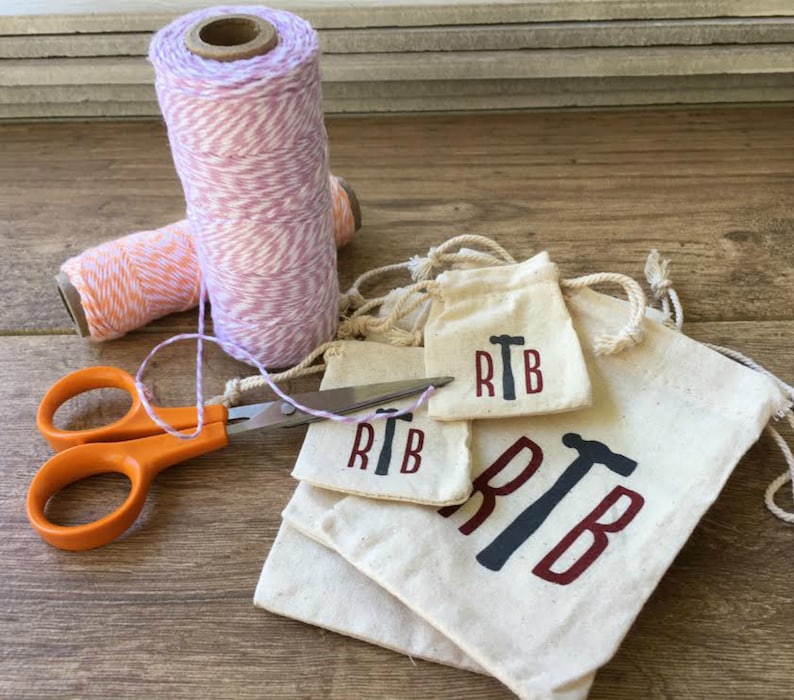 K&ouml;nnte beinhalten: Vier kleine wei&szlig;e Stoffbeutel mit Kordelzug. Die Beutel sind &uuml;bereinander gestapelt. Der gr&ouml;&szlig;te Beutel tr&auml;gt die Buchstaben "RTB" in Rot mit einem schwarzen Hammer-Motiv. Die kleineren Beutel haben das gleiche Design, sind aber kleiner. Eine orangefarbene Schere und zwei Garnrollen befinden sich im Hintergrund.