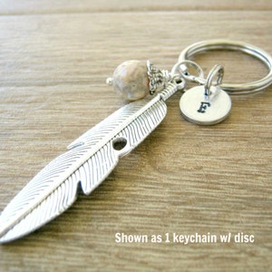 May include: Silver feather keychain with a small, round, gray and white stone charm and a silver disc charm with the letter "E" engraved on it. Shown as 1 keychain w/ disc.