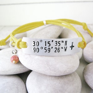 May include: A yellow suede bracelet with a silver charm engraved with the coordinates 30°15′35″N 90°59′26″W and a compass symbol. The bracelet has a pink charm attached.