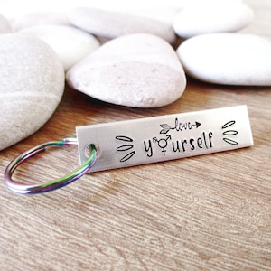 May include: A rectangular silver keychain with the words "love yourself" and gender symbols. The keychain has a rainbow-colored key ring. The text is in a handwritten style. The background is white stones.