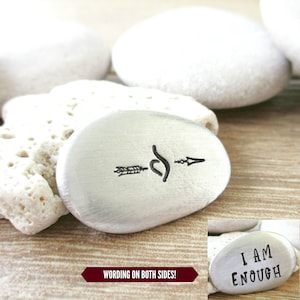May include: Silver-toned metal rock with an arrow and infinity symbol on one side and the text "I AM ENOUGH" on the other side.