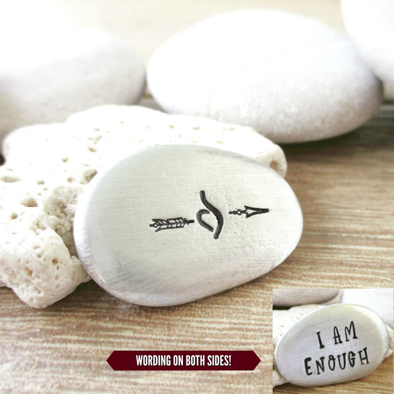 May include: Silver-toned metal rock with an arrow and infinity symbol on one side and the text "I AM ENOUGH" on the other side.