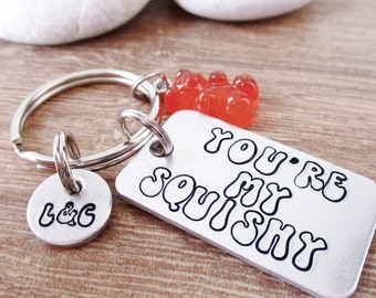 Puede incluir: Un llavero plateado con una etiqueta rectangular que dice "YOU'RE MY SQUISHY" en letras negras. Una etiqueta redonda más pequeña tiene estampado "L&G". El llavero también presenta un grupo de dijes de ositos de goma naranja.