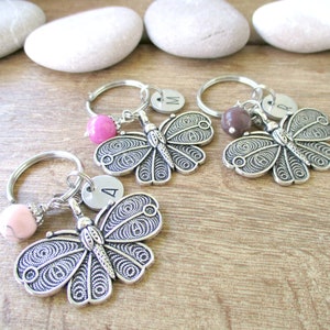 Puede incluir: Tres llaveros de mariposa plateados con cuentas rosas, marrones y blancas. Cada llavero tiene un dije plateado con una letra: A, M y R.