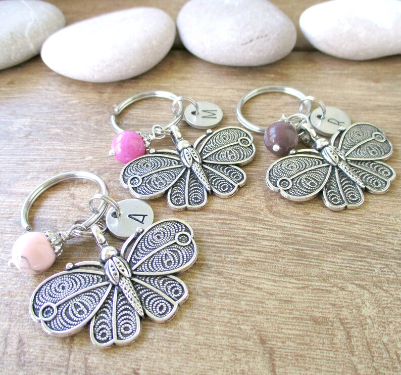 Puede incluir: Tres llaveros de mariposa plateados con cuentas rosas, marrones y blancas. Cada llavero tiene un dije plateado con una letra: A, M y R.