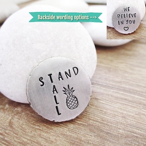 May include: A silver metal disc with the words "STAND TALL" engraved on it. A pineapple graphic is below the text. The disc is sitting on a light brown wooden surface.