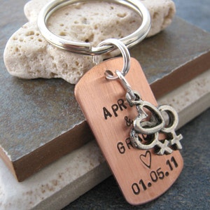 May include: Copper keychain with a silver charm of two intertwined hearts and a symbol of female gender. The keychain is engraved with the text "APR & GR 01.05.11".