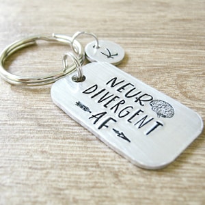 May include: Silver keychain with a tag that reads "Neuro Divergent AF" with a brain graphic. The tag is attached to a silver key ring with a small silver charm.