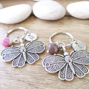 Puede incluir: Dos llaveros de mariposa plateados con un abalorio de cuentas rosa y marr&oacute;n. Cada llavero tiene un disco plateado con la letra "M" y "R" grabadas.