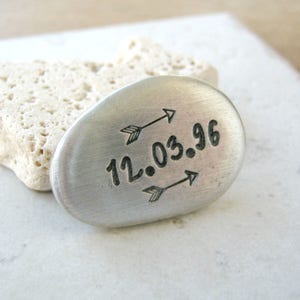 May include: A silver oval-shaped charm with the date "12.03.96" engraved on it. Two arrows point towards the date.