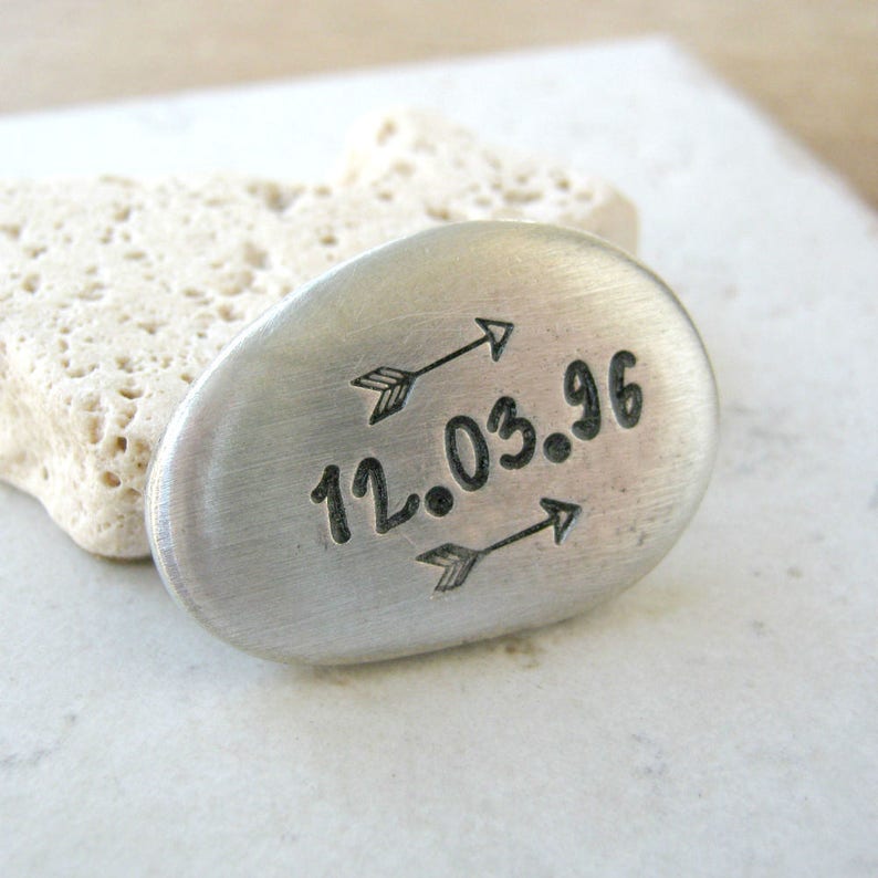 May include: A silver oval-shaped charm with the date "12.03.96" engraved on it. Two arrows point towards the date.