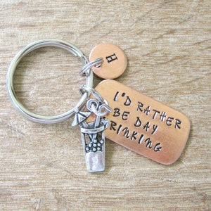 May include: A keychain with a silver ring, a copper tag with the text "I'd rather be day drinking", a small copper circle with the letter "H", and a silver charm of a cocktail. The background is a light brown wooden surface.