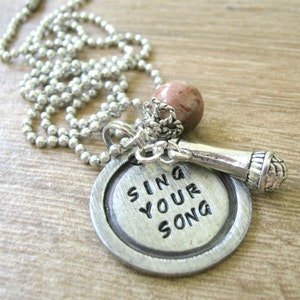 May include: A silver-toned necklace featuring a pendant with the words "SING YOUR SONG" engraved on it. The pendant is paired with a microphone charm and a small, round, brown bead. The necklace has a ball chain.