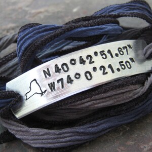 May include: A silver bracelet with a charm featuring the outline of New York State and the coordinates "N 40.642.51.67" and "W 74.0.21.50". The bracelet is made of multiple layers of blue and gray fabric.