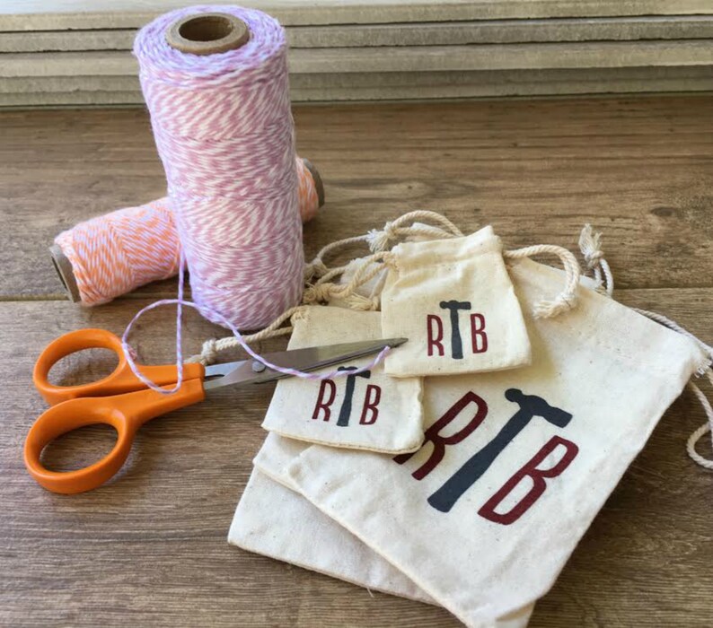 May include: Four small white fabric bags with a red and black hammer design and the initials "RTB" printed on them. The bags are stacked on top of each other. There is a pair of orange scissors and two spools of twine in the background.
