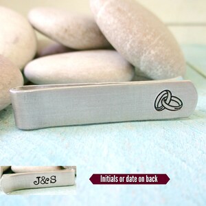 May include: Silver tie clip with two interlocking rings engraved on the front. The back of the tie clip has the initials "J&S" engraved.