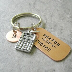 May include: A silver calculator charm, a copper charm with the text "Weapon of Choice", and a copper charm with the letter "M" are attached to a silver key ring.