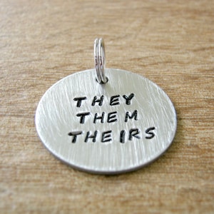 May include: A silver charm with the words "THEY THEM THEIRS" engraved on it.