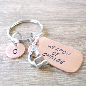 May include: A copper keychain with a silver charm and a round copper tag with the letter "C" engraved on it. The tag reads "Weapon of Choice".