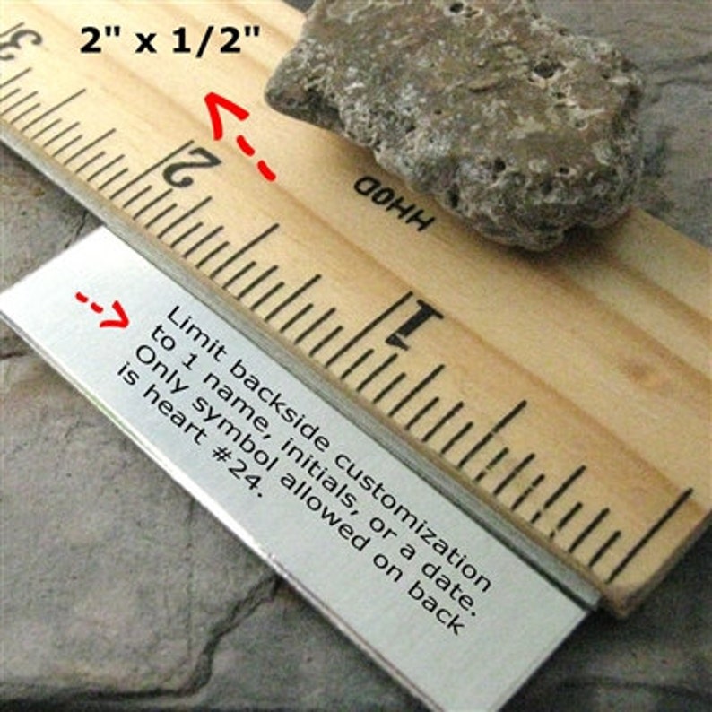 K&ouml;nnte beinhalten: Ein Holzlineal mit einem Metallstreifen, auf dem "Limit backside customization to 1 name, initials, or a date. Only symbol allowed on back is heart #24." steht. Das Lineal ist in Zoll unterteilt und zeigt eine Messung von 2 Zoll mal 1/2 Zoll.
