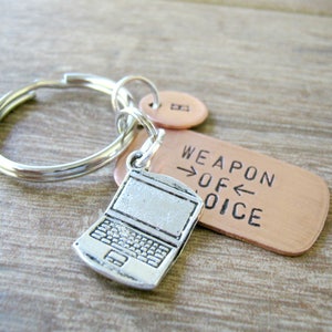May include: A silver laptop charm attached to a copper keychain with the text "Weapon of Choice" on the tag.