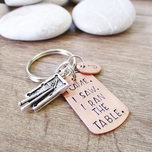 May include: A copper keychain with the text "I came, I saw, I ran the table."  The keychain also features a silver charm of a pool table with cues.