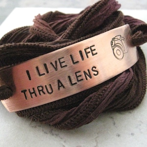 May include: A copper bracelet with the words "I live life thru a lens" and a small camera icon engraved on it. The bracelet is wrapped in a brown fabric.