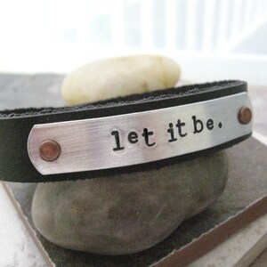 May include: A black leather bracelet with a silver metal plate that says "let it be." The plate has two copper rivets on either side.