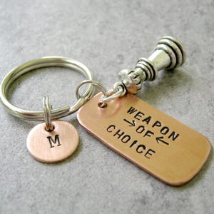 May include: A copper keychain with a silver chess piece charm and a tag that reads "Weapon of Choice". The keychain also has a small copper disc with the letter "M" engraved on it.