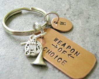 Puede incluir: Un llavero de cobre con un dije de trompa francesa de plata. El llavero tiene una etiqueta que dice "Weapon of Choice". También hay una pequeña etiqueta de cobre con la letra "M" en ella.