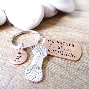 May include: Copper keychain with a silver yarn charm and a copper charm with the letter K. The copper charm has the text "I'd rather be crocheting".