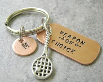 May include: A silver key chain with a copper tag that says "Weapon of Choice" and a silver tennis racket charm. There is also a copper charm with the letter "M" on it.