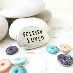 May include: A silver metal rock with the words "FOREVER LOVED" engraved on it. The rock is surrounded by small, colorful stones.