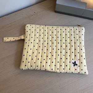 May include: A rectangular quilted pouch in pale yellow with a repeating pattern of small dark brown polka dots. The pouch features vertical stitching, a side wrist strap, a gold-toned zip, and a small white fabric label with a black flower icon.