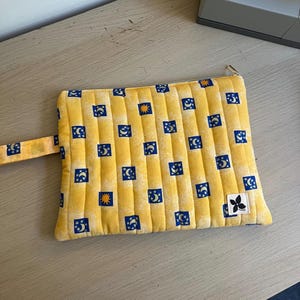 May include: A rectangular quilted zip pouch in yellow fabric featuring a repeating pattern of small blue squares containing white crescent moons, stars, and yellow suns. The pouch includes a matching yellow wrist strap and a small white label.