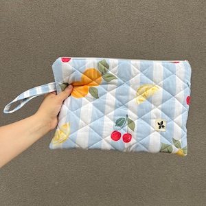 Pode incluir: Bolsa de mão acolchoada com um padrão de listras azuis e brancas, apresentando ilustrações de limões, laranjas e cerejas. A bolsa tem fecho de zíper e uma pequena alça. Um pequeno emblema de flor preta está no canto inferior direito.