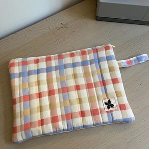 May include: A rectangular quilted pouch featuring a multi-coloured plaid pattern in shades of coral, light blue, and mustard yellow on a cream background. The pouch includes a matching wrist strap, a gold-tone zip, and a small white fabric label.