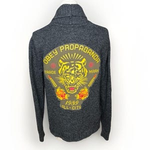 Puede incluir: Cárdigan de punto gris oscuro con cuello chal. La parte trasera presenta un parche grande con las palabras "OBEY PROPAGANDA" y un gráfico de tigre en amarillo, rojo y verde oliva. Debajo del gráfico está el texto "1989 ALL-CITY".