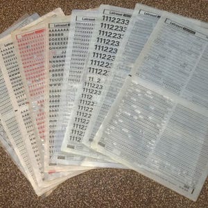 Peut inclure: Une pile de feuilles Letraset avec différentes polices de caractères et des numéros imprimés dessus. Les feuilles sont disposées en éventail, la feuille du dessus montrant les numéros 1112233, 1112233, 1111122, 111223, 111223, 111223, 11122:, 11122, 11 2, 111223, 111223, 1112, 11122, 11122, 11122, 11122, 1112, 1112.