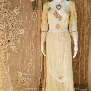 May include: A long, ivory-colored gown with delicate lace detailing and floral embroidery. The gown has a sweetheart neckline and a fitted bodice. The skirt is full and flowing, with a layer of lace at the hem. The gown is accessorized with a gold heart pendant necklace.