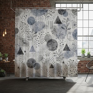 May include: A shower curtain with a watercolor design featuring gray and blue geometric shapes, including triangles, circles, and dots, along with black line art of leaves and stems. The curtain hangs in a bathroom with a brick wall.