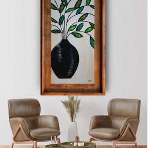 May include: Abstract painting of a black vase with green and yellow leaves in a wooden frame. The vase has a textured surface. The artwork is displayed on a white wall above two brown leather armchairs.
