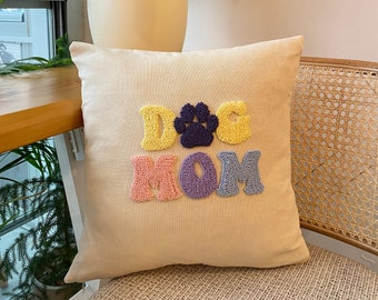 May include: A beige cushion with the words "DOG MOM" embroidered in colourful yarn. The letters are puffy and the "O" in MOM is replaced with a purple paw print.