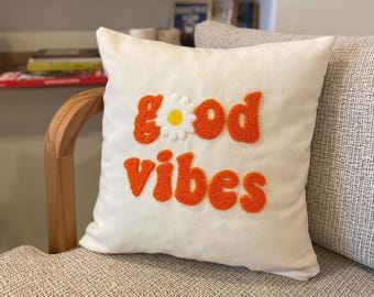May include: White cushion with the words "good vibes" in orange fluffy lettering and a white daisy with a yellow centre.