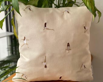 May include: A cream-coloured throw pillow with embroidered images of people doing yoga poses.