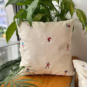 Può includere: Cuscino quadrato color crema con ricami di sciatori in varie pose. Gli sciatori indossano abiti rossi, blu, viola e gialli. Il cuscino è posto su una superficie di legno, con una pianta verde sullo sfondo.