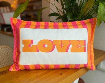 May include: Rectangular cushion featuring the word "LOVE" in orange and pink block letters on a white background. The cushion has a pink and orange striped border.