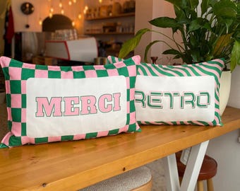 May include: Two rectangular cushions with a retro design. One cushion has the word "MERCI" in pink block letters, and the other says "RETRO". Both cushions have a white centre and a green and pink checkered or striped border.