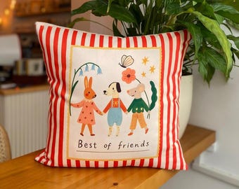 May include: Square cushion with a red and white striped border. The centre features a whimsical illustration of a rabbit, dog, and mouse holding hands, with the text "Best of friends" below.